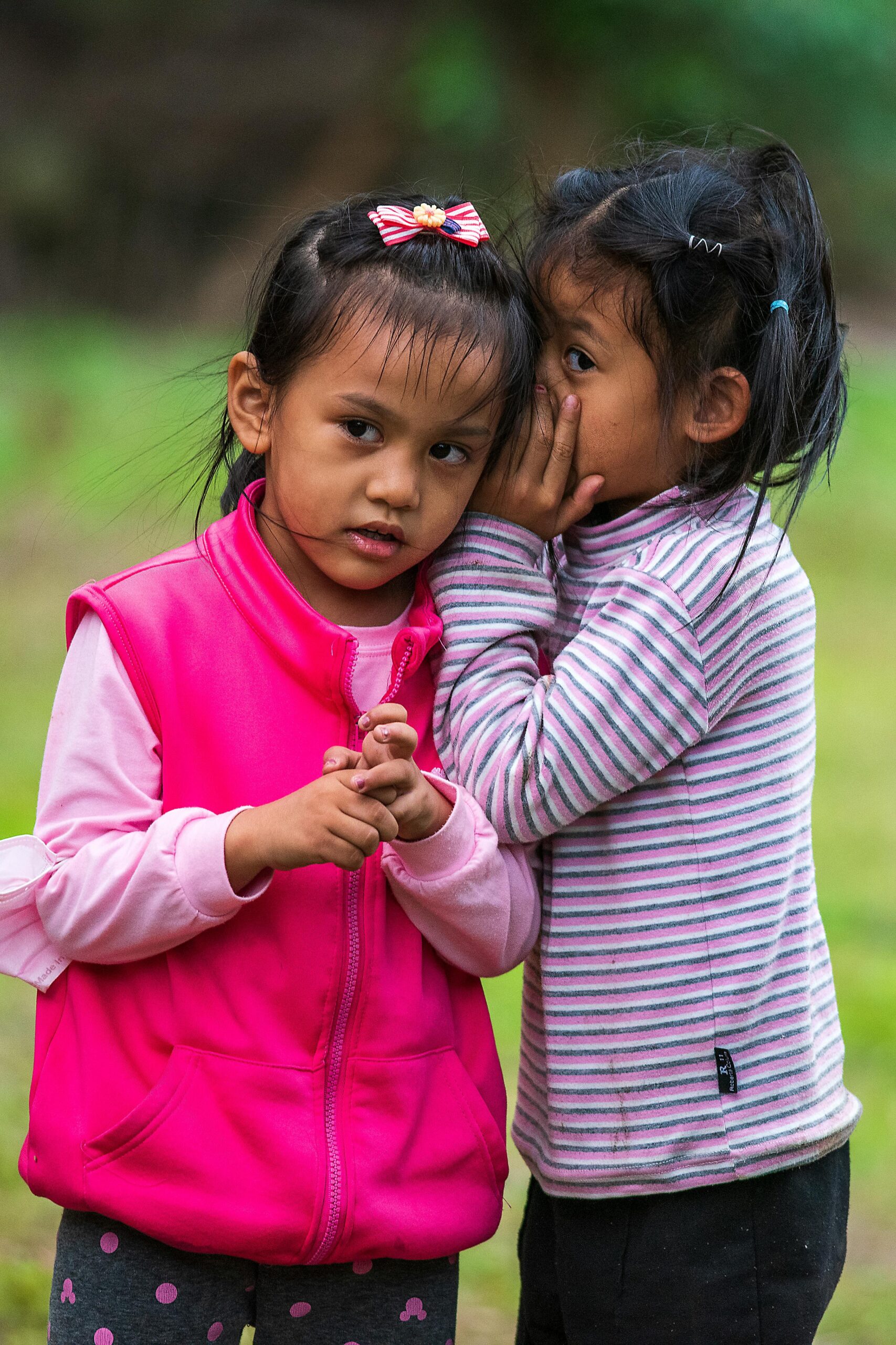 Two young girls share a secret outdoors, capturing a candid childhood moment full of innocence and friendship.